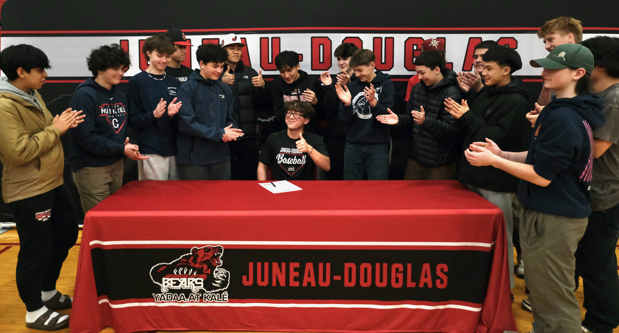 Teammates and friends applaud after Juneau-Douglas High School: Yadaa.at Kalé senior Lamar Blatnick signs a letter of intent Wednesday in the JDHS gymnasium to play baseball for Dallas Christian College. (Klas Stolpe for the Juneau Empire)