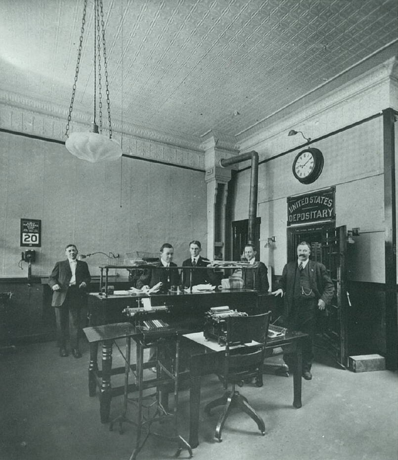 Men stand inside the First National Bank with its vault door open in 1916. In 1925 the bank moved a block away after water damage from a fire in a second floor storage room prompted the bank to relocate. Note the decorative tin ceiling, wall calendar and adjacent telephone. (Photo courtesy First National Bank Alaska)