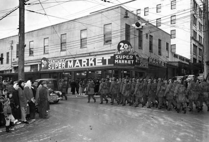 Uniformed Alaska National Guard troops carrying rifles march past the 20th Century Super Market on a rainy day in about 1959 as spectators watch from across the street. Note the bland stripped down building exterior that seems to match the dreary weather. (Photo credit ASL-Alaska-National-Guard-18)