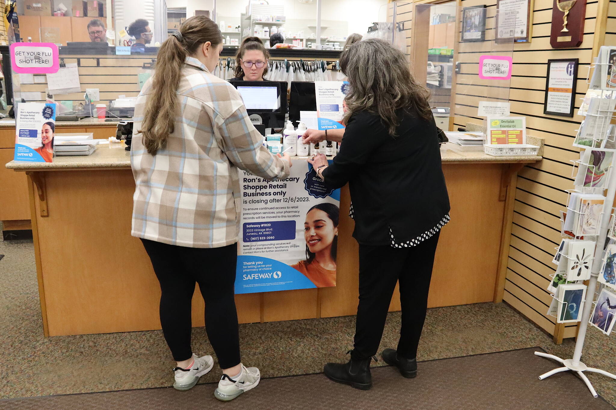 Cheyenne Latu (left), a pharmacy technician at Ron’s Apothecary Shoppe, and business co-owner Gretchen Watts hang a poster at the front counter Thursday announcing the store’s closure after Dec. 6 as Jessica Kirtley, another pharmacy technician, works at the front register. The nearby Safeway supermarket has agreed to take the prescriptions of all customers as well as hire all of the independent pharmacy’s employees, according to the co-owners who are retiring. (Mark Sabbatini / Juneau Empire)