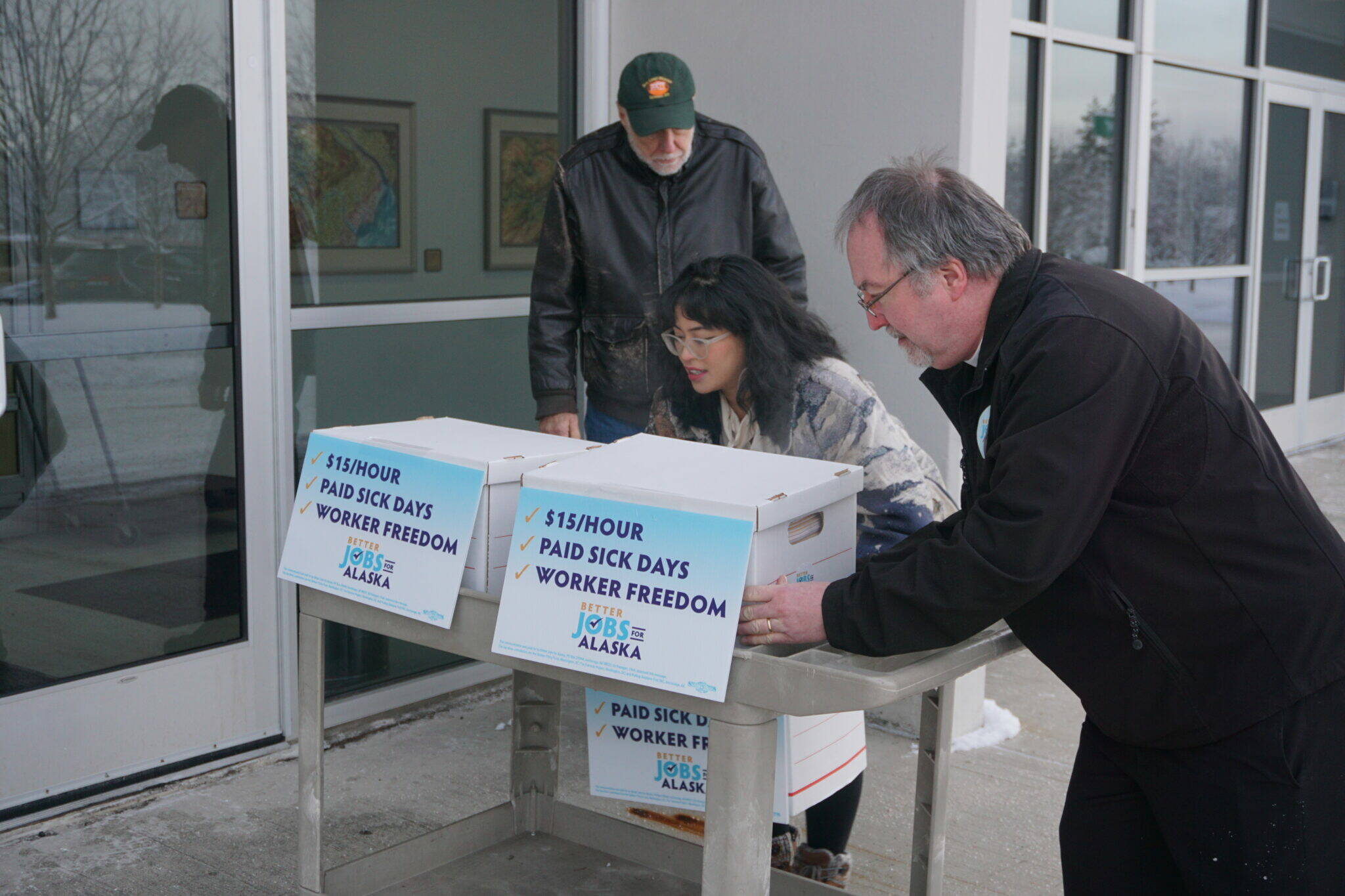 Former state labor commissioner Ed Flanagan, State Rep. Genevieve Mina, D-Anchorage, and the Rev. Michael Burke of St. Mary’s Episcopal Church in Anchorage wheel boxes of signed petitions into a state Division of Elections office on Jan. 9. The petitions were for a ballot initiative to increase the state’s minimum wage, mandate paid sick leave and ensure that workers are not required to hear employers’ political or religious messages. (Photo by Yereth Rosen/Alaska Beacon)