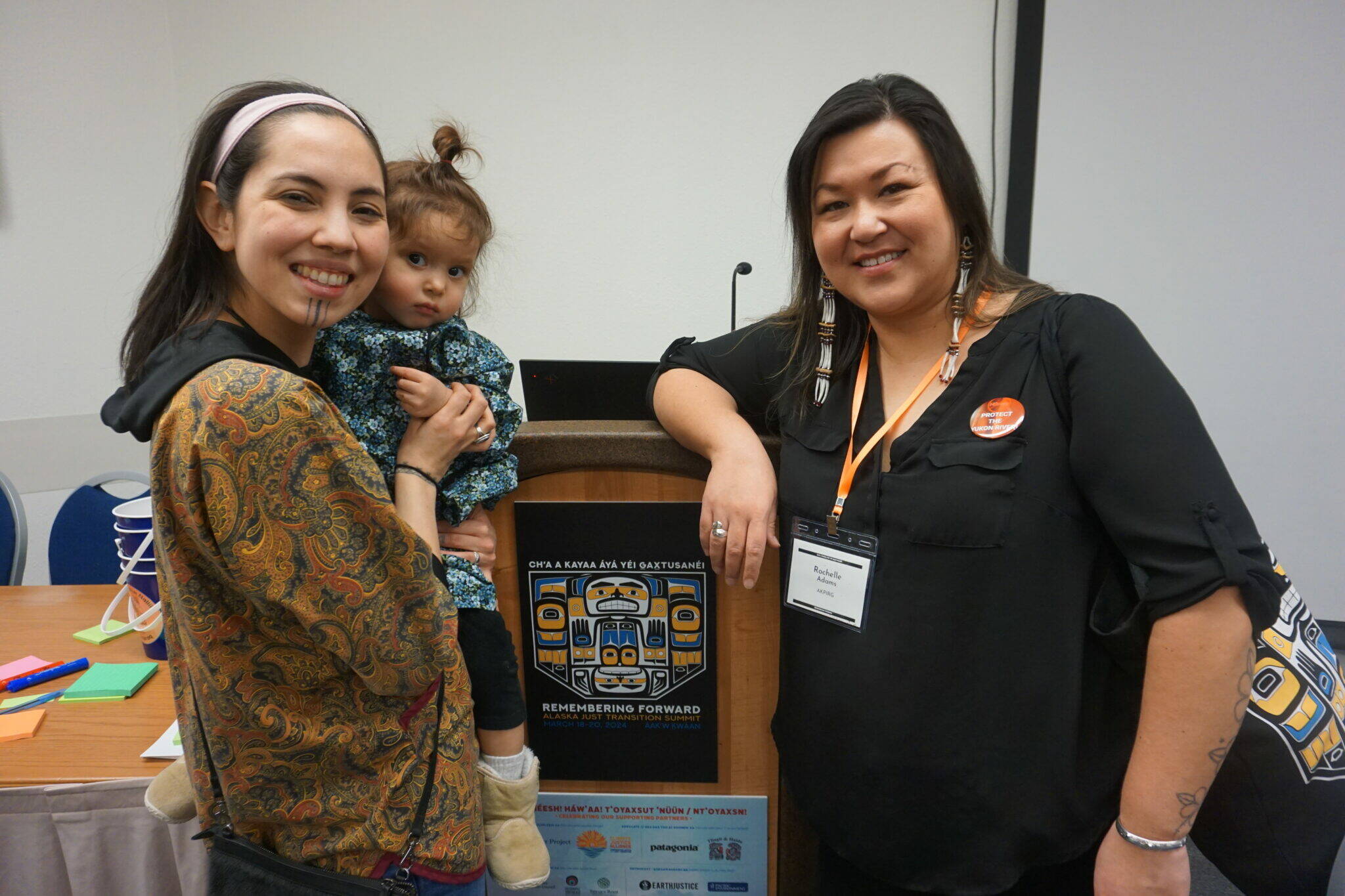 Annauk Olin, holding her daugher Tulġuna T’aas Olin, and Rochelle Adams pose on March 20, 2024, after giving a presentation on language at the Alaska Just Transition Summit in Juneau. The two, who work together at the Alaska Public Interest Research Group’s Language Access program, hope to compile an Indigenous environmental glossary. (Photo by Yereth Rosen/Alaska Beacon)