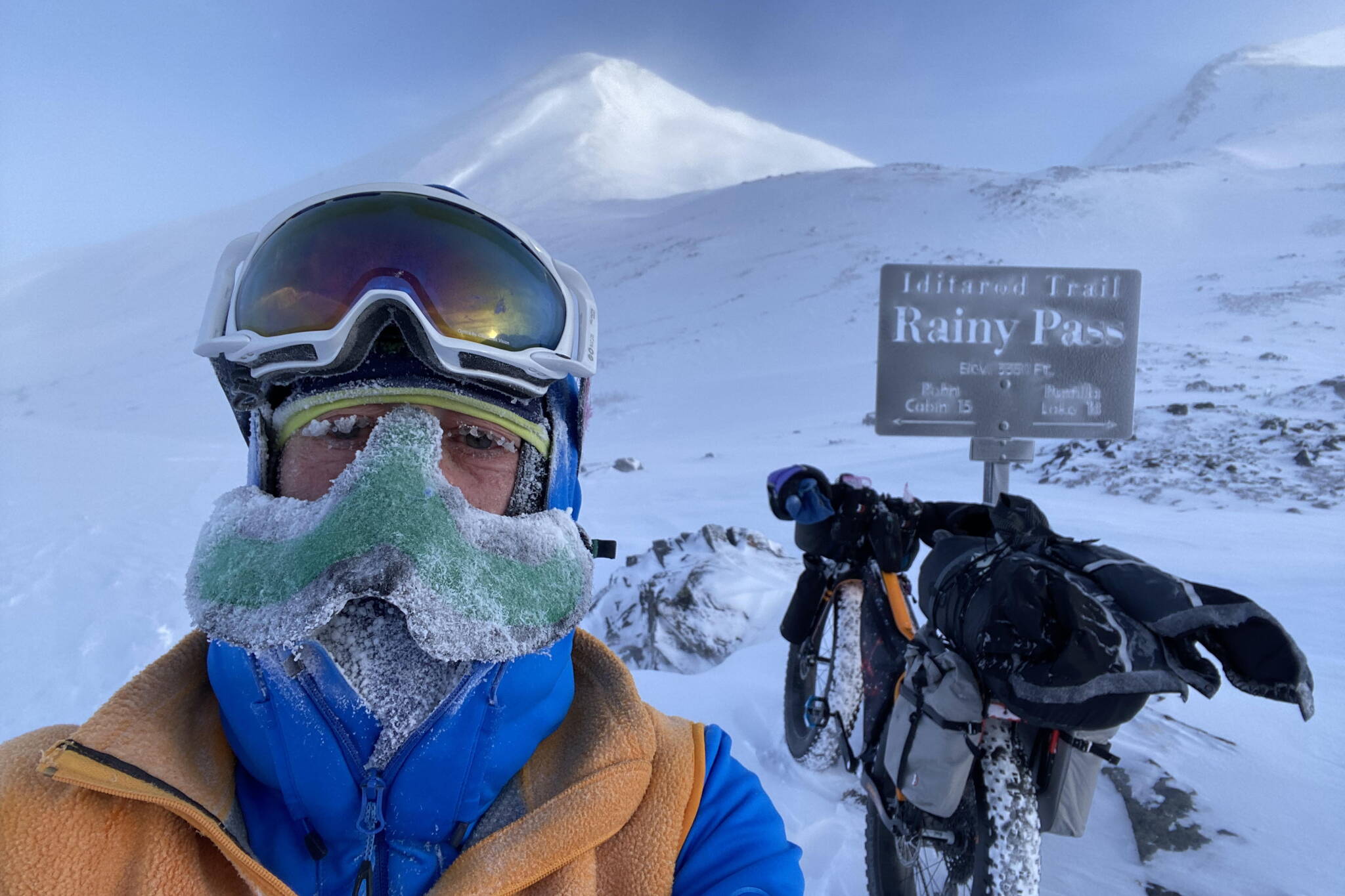Peter Delamere poses in front of a sign in Rainy Pass in the Alaska Range during the 2024 Iditarod Trail Invitational race. On his face is a “nose-hat” invented by Fairbanks athlete Shalane Frost. (Photo by Peter Delamere)