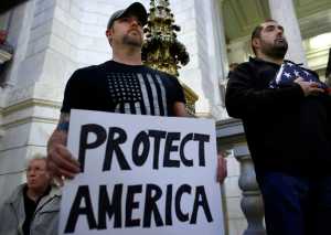 U.S. Army veteran Jim Purcell, of Burrillville, R.I., front left, displays a placard as U.S. Navy veteran Robert Martinez, right, displays a folded American flag during a rally Thursday, Nov. 19, 2015, at the Statehouse, in Providence, R.I., held to demonstrate against allowing Syrian refugees to enter Rhode Island following the terror attacks in Paris. (AP Photo/Steven Senne)