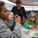 IMAGE DISTRIBUTED FOR MATTEL, UNO - Former Mayor and current Assembly member Mary Becker watches as Juneau Community Charter School students Clinton McQueen, 11, left, Brynna Morgan, 8, center, and Lamar Blatnick, 9, play a game of UNO in the Assembly Chambers in Juneau, Alaska, on Friday, April 1, 2016. As part of an April Fools' promotion, the city of Juneau and Mattel announced a renaming of Juneau to UNO. (Michael Penn/AP Images for Mattel, UNO)