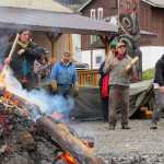 In the foreground, from left, carvers Zack James, Ted Hart and James Hart dance as Ted Hart drums. Zack James and James Hart are in Hoonah from Haines for several months to carve two forty-foot spruce canoes for the ceremonial return of the Huna Tlingit to Glacier Bay; Ted Hart came down with his family for the week to help out. In the foreground is the fire on which they're heating the rocks to steam the canoe, covered with a tarp in the background.