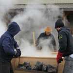 Don Bolton, left, and James Hart carry fire-heated rocks to the canoe for steaming as lead carver Wayne Price evaluates the expansion's progress.