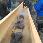 Carvers and community members place hot rocks in the canoe as the steaming process begins. Others wait to cover the canoe with a tarp.