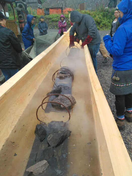 Carvers and community members place hot rocks in the canoe as the steaming process begins. Others wait to cover the canoe with a tarp.