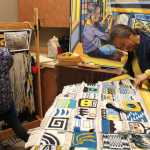 Mother and daughter - weaver Lily Hope, left, works on a robe (backdropped by an Arnie Weimer painting of her working on the same project) as her mother, Clarissa Rizal, works on the "Weavers Across the Waters" robe 40 weavers from around the Pacific Northwest contributed to. It will be used for canoe gatherings and the maiden launch of new dugout canoes.