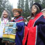 From left, weavers Marsha Hotch, Clarissa Rizal, and Irene Lampe watch as canoers proceed, with one of the dugout canoes, up the beach. Rizal is holding the Weavers Across the Waters robe whose creation she spearheaded. Forty-four weavers from up and down the Northwest Coast contributed to the robe, including each of these three women.