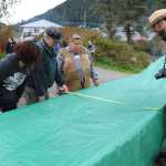 Those whose faces are visible are, clockwise from left, lead carver Steve Brown, with tape measure as he measures the expanded width of the canoe, Shangukeidí (Thunderbird) clan leader David Katzeek, and Sitka National Historical Park  acting chief of interpretation Ryan Carpenter.