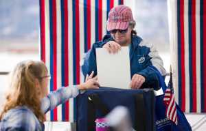Election official Kim Peterson, left, helps voter Diana Quinones drop her ballot into the box after voting early at the State Office Building on Monday. Early voting can also be done at the Mendenhall Mall.