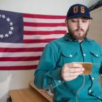 In front of a replica 1776 U.S. flag, KC Mack hand sews a wallet from the leather of a used baseball mitt on Friday, Nov. 17, 2017. (Michael Penn | Juneau Empire)