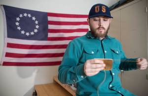 In front of a replica 1776 U.S. flag, KC Mack hand sews a wallet from the leather of a used baseball mitt on Friday, Nov. 17, 2017. (Michael Penn | Juneau Empire)