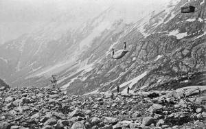 Long Hill, Chilkoot Trail, looking southwest. This view shows the Chilkoot Railroad & Transport Company (CR&T) aerial tramway in operation. One of the CR&T tramway towers is in the background left and a canoe and crate are being hauled over the line toward the summit of the Chilkoot Trail. Two men, possibly tramway workers, are observing the load. This photograph was taken on Long Hill between Sheep Camp and the Scales between the spring-fall of 1898-1899. Image courtesy of the National Park Service, Klondike Gold Rush National Historical Park, Candy Waugaman Collection, KLGO LH-68-8973.
