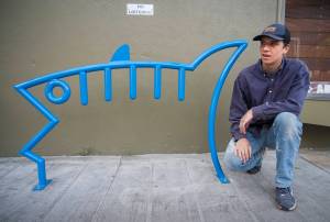 Ben Campbell, a recent graduate of Juneau-Douglas High School, kneels next to one of two of his fish designed bicycle racks on Ferry Way on Wednesday, June 13, 2018. Campbell designed the rack for a class and the city choose to make it a reality. (Michael Penn | Juneau Empire)