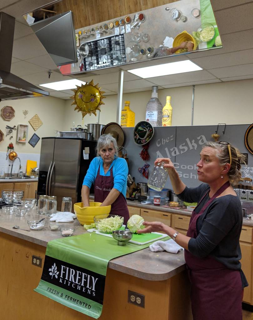 Julie OBrien, a University of Alaska Southeast alumna and owner of Firefly Kitchens, holds a jar up while Julie Hamilton, a assistant professor of accounting for UAS, works on cut cabbage during Fermentation 101. The workshop focused on the benefits of fermented foods and how folks could make their own. The large mirror above the counter at Chez Alaska Cooking School allowed the audience to see how the food prep was done. (Ben Hohenstatt | Capital City Weekly)