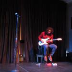 Avery Stewart on stage during the Woosh Kinaadeiyí Poetry Slam, Oct. 20. With the pedals near his feet, Stewart can provide backing instrumental loops for his guitar playing. (Ben Hohenstatt | Capital City Weekly)