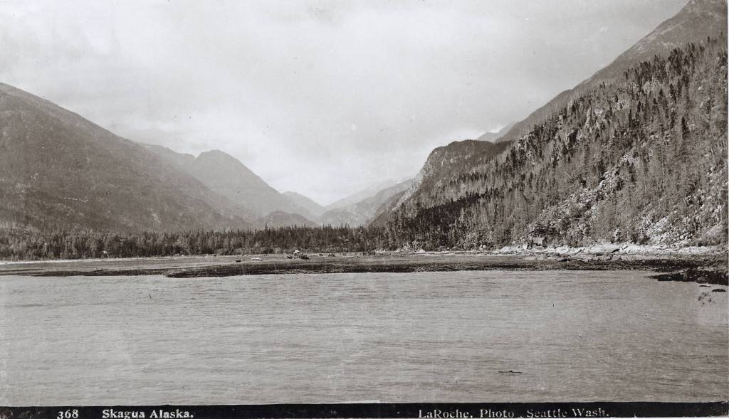 This photograph of the Skagway valley was taken from the deck of the Steamer Queen on July 26, 1897 and shows the almost undisturbed terrain of the Skagway Valley just before the Klondike Gold Rush begins for Skagway. This is Mooresville, Captain Moores name for his little hamlet. In the next few hours, days, weeks, and months, the gold seeking Argonauts will invade the valley like an army with their outfits, tents and animals. In less than a month, a street grid will be laid out. The tents of the first month will quickly give way to wood buildings, one, two, and three stories high and the population will increase from around two dozen or so to over 10,000 by the start of 1898. In the center of this image stand Captain William Moores Cabin from 1887 and in front, his son Ben Moores wood-frame house under construction. (Library of Congress, 3c22304u; KLGO SE-9-8799)