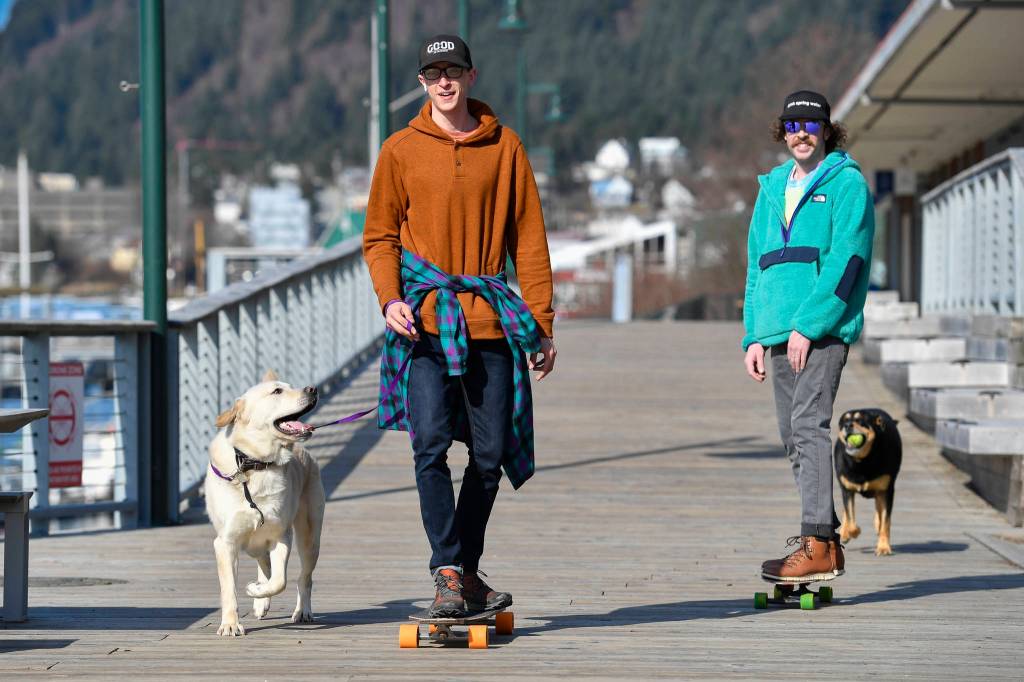 Cody Kopkie, left, with Olly, and Ray Huebschen, followed by Nala, take advantage of the sunny weather and lack of ice to skateboard along the Seawalk on Monday, March 25, 2019. (Michael Penn | Juneau Empire)