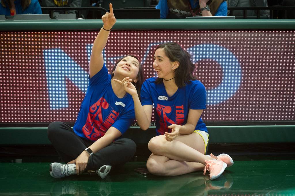 Team Juneaus Sara Steeves and Kaytlynne Lewis view a video in front of the Native Youth Olympics logo at the Alaska Airlines Center in Anchorage, Alaska, on Thursday, April 25, 2019. (Michael Dinneen | For the Juneau Empire)