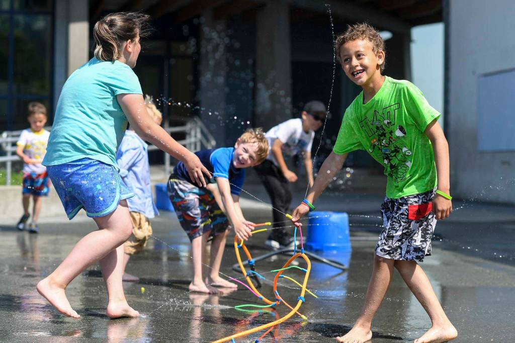 David Kimbrough, 7, right, Clayton Haywood, 6, center, and Kyla Belcourt, 8, play in sprinklers set up during the RALLY program at Harborview Elementary School on Wednesday, June 26, 2019. (Michael Penn | Juneau Empire)