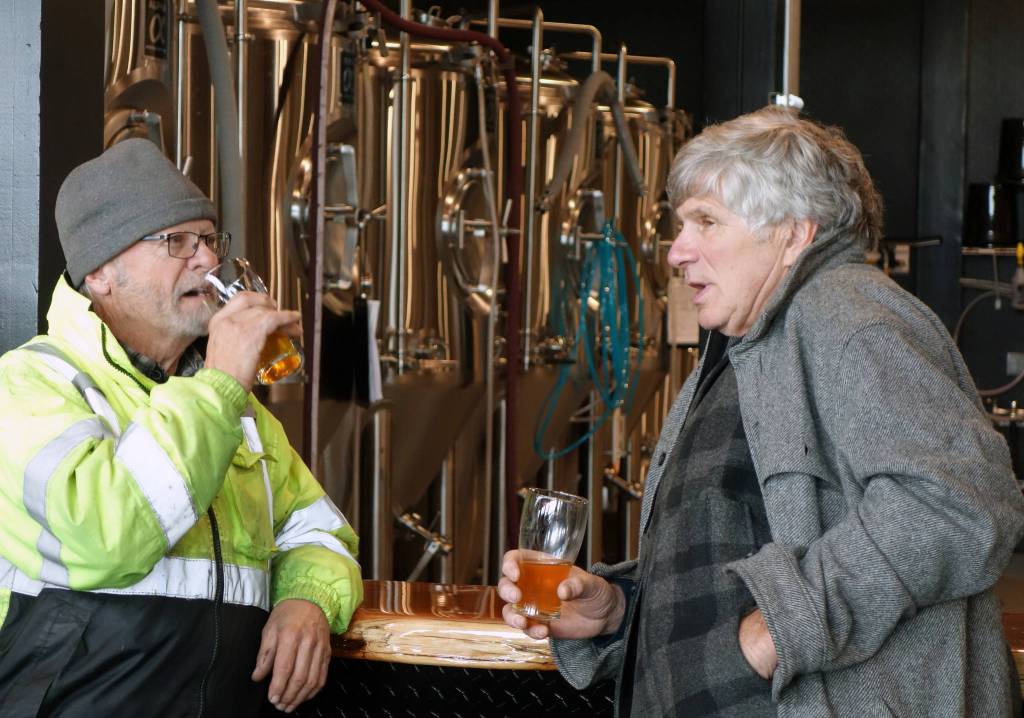 Dave Iverson and Bill Ashby enjoy beers and conversation in front of brewery equipment at Forbidden Peak Brewerys grand opening, Saturday, Oct. 12, 2019. (Ben Hohenstatt | Juneau Empire)