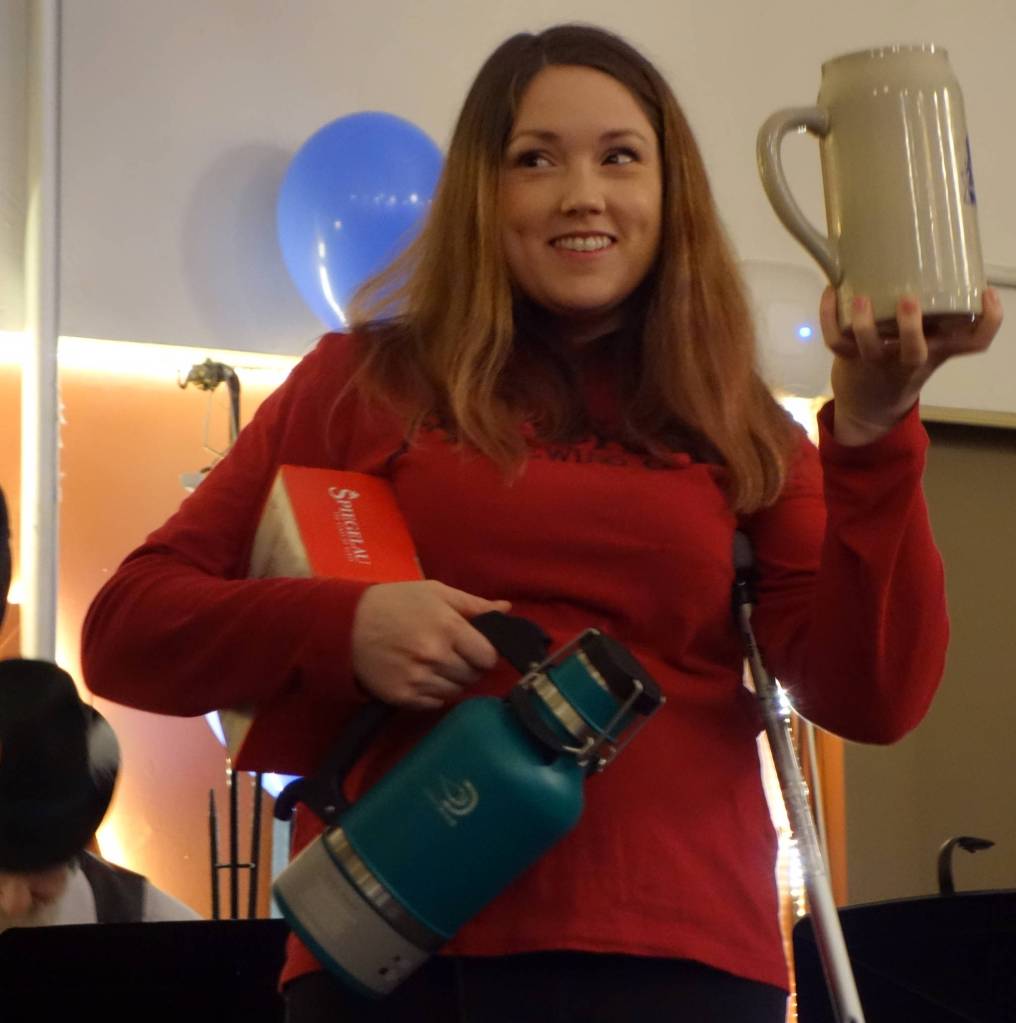 Womens strong stein contest winner Maureen Shivers clutches her winnings in one arm, hoists her stein with another and balances on a crutch at AKtoberfest, Saturday, Oct. 12, 2019. (Ben Hohenstatt | Juneau Empire)