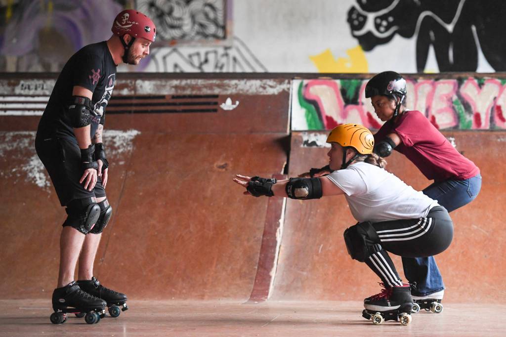 Jennifer Gross, right, and Shabadrang Khalsa, who skate with the Juneau Rollergirls, teach Empire reporter Michael S. Lockett how to rollerskate at the Pipeline Skate Park on Friday, Oct. 11, 2019. (Michael Penn | Juneau Empire)