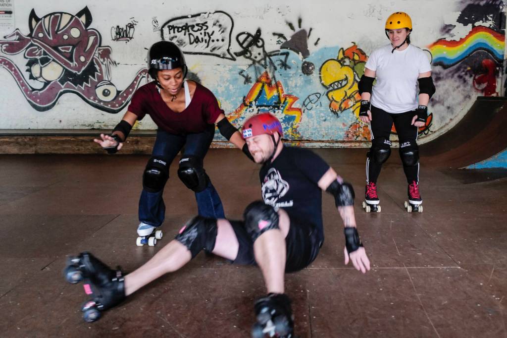 Empire reporter Michael S. Lockett takes a fall while being taught to rollerskate by Jennifer Gross, right, and Shabadrang Khalsa, at the Pipeline Skate Park on Friday, Oct. 11, 2019. (Michael Penn | Juneau Empire)