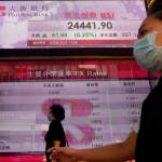 People wearing face masks walk past a bank electronic board showing the Hong Kong share index at Hong Kong Stock Exchange Monday, April 20, 2020. Shares were mixed in Asia on Monday, while oil prices have fallen back. (AP Photo | Vincent Yu)