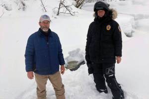This photo provided by the Alaska Wildlife Troopers taken March 9, 2021, Doug Ramsey, left, of Sundance Wyoming, poses with Alaska Wildlife Trooper Jason Kneier near a hole in the ice of a river in Swentna, Alaska. The two helped pull an 8-year-old boy from the water after he fell into the river. (Alaska Wildlife Troopers)