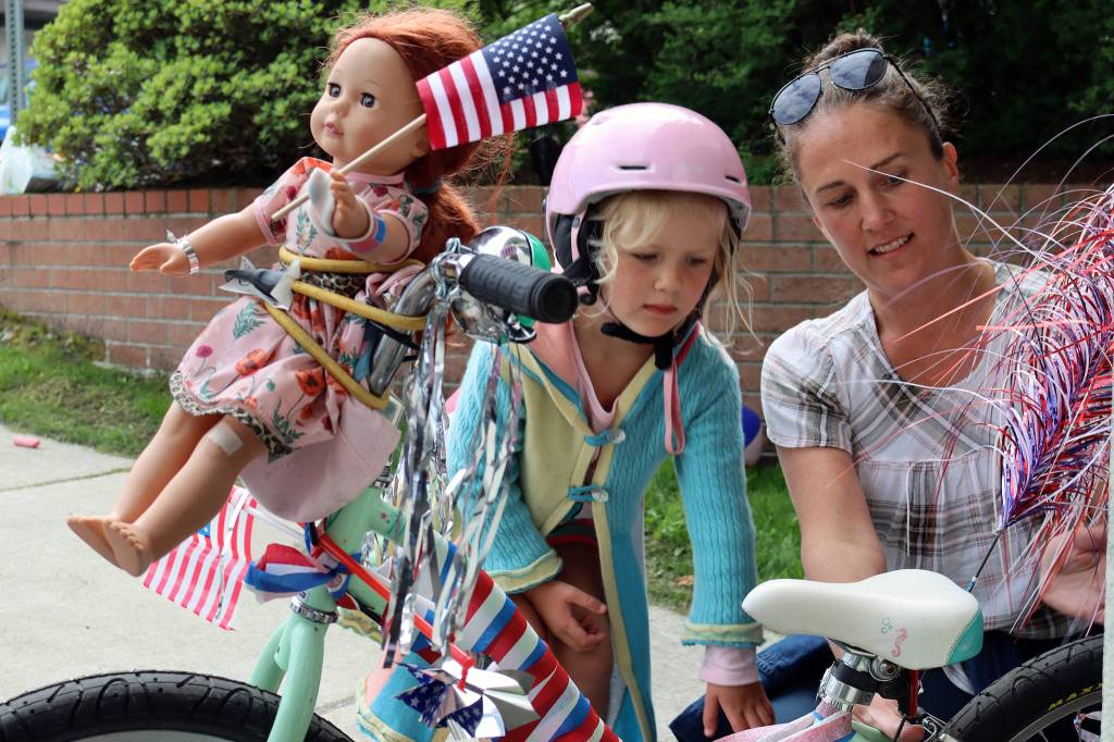 Imogen Resneck, 4, and Jamie Buehner finish up decorating a bike outside the Douglas Public Library on July 3, 2021. Resnecks doll, Robbie, was bound to the bike like an amber-haired figurehead because she wanted to go to the parade, Resneck said. (Ben Hohenstatt / Juneau Empire)