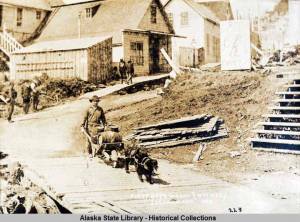 This photo from the Capt. George H. Whitney Photograph Collection shows a man, with wheelbarrow cart and two dogs in harness, transports beer barrels along boardwalk; pedestrians, buildings, and sign for Coons Drugstore in background in Juneau in 1886. Juneau and Douglas breweries were the subject of the Gastineau Channel Historical Societys award-winning newsletter. (William Howard Case/ Alaska State Library - Historical Collections)