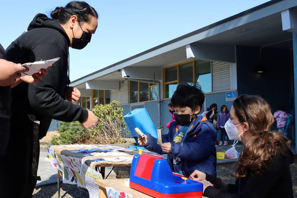 Aiona Fisi gives Daniel Lee, a kindergarten student, money for a card decorated with a student-drawn picture of a sunflower, while kindergarten teacher Carly Lehnhart supervises. (Ben Hohenstatt / Juneau Empire)