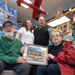 The three generations of Wileys who have operated the Ben Franklin Store are shown inside the shop. Seated are Fred and Sally Wiley, holding a painting of the 1930s Blomgren Building. Standing are Meagan Wiley Bishop and her father Mike Wiley who currently run the popular downtown Front Street business. (Michael Penn / Downtown Business Association)