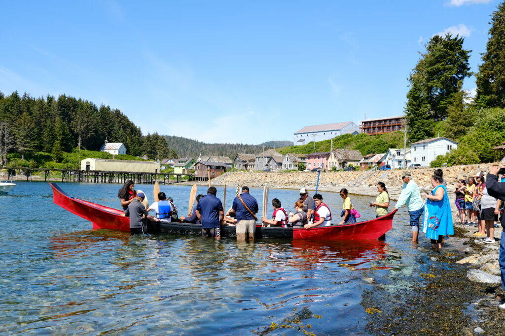 Angoon students prepare to paddle the unity canoe they built with master carver Wayne Price. Crew from the Polynesian voyaging canoe Hōkūle‘a help steady the canoe while their teacher, Chenara Johnson, looks on June 19. (Photo by Claire Stremple/Alaska Beacon)