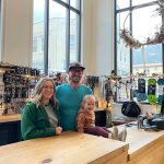 Foggy Mountain Shop owners Sean Reilly and Courtney Nicholl with their daughter Alder stand at their custom-built Icy Straits Lumber counter. Courtney designed the space to focus attention on inspiring outdoors activities with the steep mountain backdrop. (Photo by Laurie Craig)