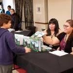Felicia Price, an employee of the Central Council of Tlingit and Haida Indian Tribes of Alaska, hands a copy of the Lingít-language book Kuhaantí to her son, Brayden, 8, while staffing the distribution table for the book with co-worker Genevieve McFadden during its release party Friday night at Elizabeth Peratrovich Hall. (Mark Sabbatini / Juneau Empire)