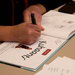 G̱at X̱wéech Nick Alan Foote signs a copy of the Lingít-language book Kuhaantí he helped illustrate during a release event Friday night at Elizabeth Peratrovich Hall. (Mark Sabbatini / Juneau Empire)