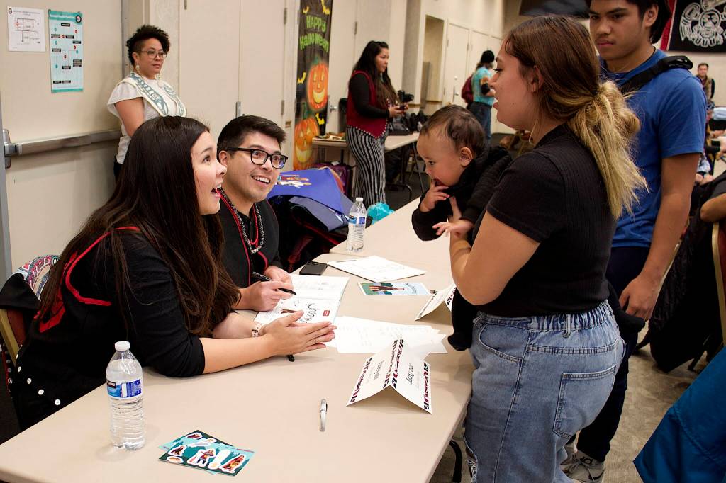 Jaax̱snée Kelsey Mata Foote (left) and G̱at X̱wéech Nick Alan Foote, illustrators of the Lingít-language book Kuhaantí, talk to Kapualani Bagoyo, her nine-month-old daughter Lashya, and Iverson Niuatoa at a signing table during the books launch Friday night at Elizabeth Peratrovich Hall. (Mark Sabbatini / Juneau Empire)