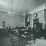 Men stand inside the First National Bank with its vault door open in 1916. In 1925 the bank moved a block away after water damage from a fire in a second floor storage room prompted the bank to relocate. Note the decorative tin ceiling, wall calendar and adjacent telephone. (Photo courtesy First National Bank Alaska)