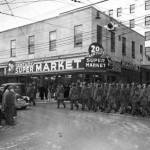 Uniformed Alaska National Guard troops carrying rifles march past the 20th Century Super Market on a rainy day in about 1959 as spectators watch from across the street. Note the bland stripped down building exterior that seems to match the dreary weather. (Photo credit ASL-Alaska-National-Guard-18)