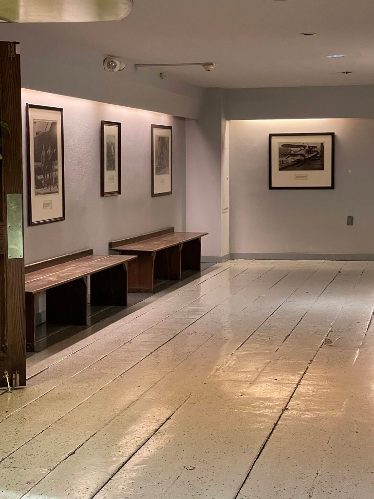 The original 1939 hangars wood plank decking is preserved under gray paint in the corridor of Merchants Wharf. On the walls hang framed sepia-toned historic aviation photos with captions. (Photo Laurie Craig)