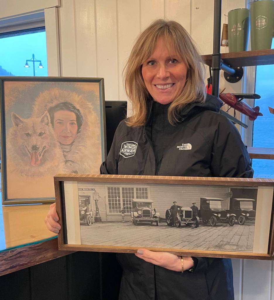 Wings Airways co-owner Holly Johnson displays a photo of the Johnson familys 1920s Ford dealership on the site of todays Merchants Wharf. Behind her is a painting of Juneau dog mushing celebrity Mary Joyce who owned Taku Lodge in the 1930s. Wings Airways partners with current lodge owners Ken and Michelle Ward who provide wild salmon meals for flightseeing guests in summer. (Photo Laurie Craig)