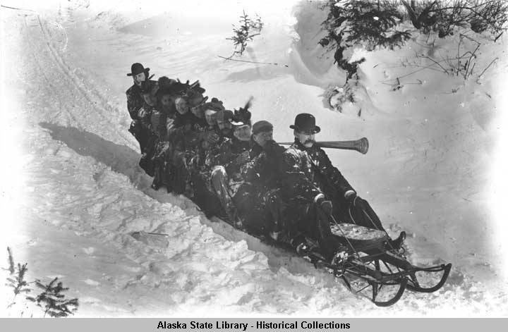 Ten adults pack onto a long sled to slide down a snow-packed slope. Often city streets were used for this winter activity. One man holds a horn, ready to warn pedestrians of the oncoming sledders. (Alaska State Library  Historical Collections P87-1258)
