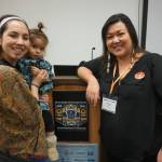 Annauk Olin, holding her daugher Tulġuna Taas Olin, and Rochelle Adams pose on March 20, 2024, after giving a presentation on language at the Alaska Just Transition Summit in Juneau. The two, who work together at the Alaska Public Interest Research Groups Language Access program, hope to compile an Indigenous environmental glossary. (Photo by Yereth Rosen/Alaska Beacon)