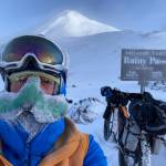 Peter Delamere poses in front of a sign in Rainy Pass in the Alaska Range during the 2024 Iditarod Trail Invitational race. On his face is a nose-hat invented by Fairbanks athlete Shalane Frost. (Photo by Peter Delamere)