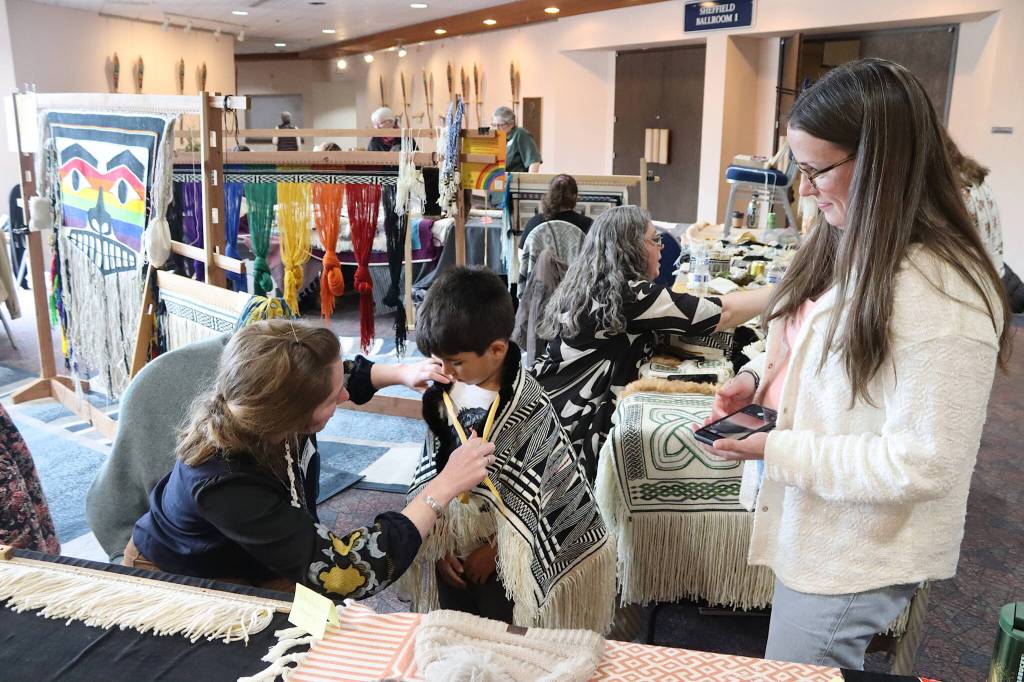 Stephanie Hayes (left), a Petersburg resident, puts a Ravenstail robe on Preston Singletary while his mother Michelle watches during a weavers gathering preceding a ceremony featuring the robes Tuesday at Centennial Hall. (Mark Sabbatini / Juneau Empire)